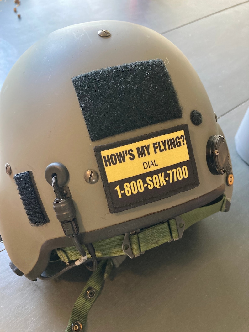 May include: A green military helmet with a black velcro patch and a yellow sticker that reads "How's My Flying? Dial 1-800-SQK-7700".