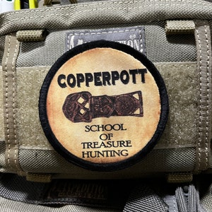 Op de afbeelding: Een beige en zwarte geborduurde patch met de tekst "COPPERPOTT SCHOOL OF TREASURE HUNTING" en een gestileerde afbeelding van een sleutel.