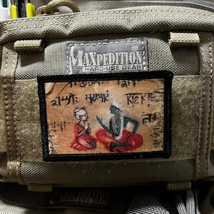 May include: A Maxpedition Hard-Use Gear bag with a patch featuring a colourful illustration of two figures in traditional clothing. The patch has text in a foreign language.