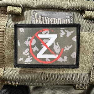 May include: A black and gray patch with a white letter Z with a red circle and a diagonal line through it. The patch has a camouflage pattern and the text "MAXPEDITION" is visible on the bag.