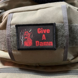 Puede incluir: Un parche negro con texto rojo que dice "Give A Damn". Un diablo de dibujos animados rojo con cuernos y un tridente está en la esquina superior izquierda del parche.