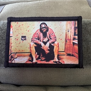May include: A black and white embroidered patch featuring a man sitting on the floor in a room with a yellow and red patterned wallpaper. The man is wearing a red robe, black pants, and sunglasses.