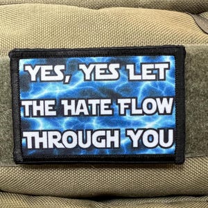 Puede incluir: Un parche rectangular con el texto "YES, YES LET THE HATE FLOW THROUGH YOU" en letras blancas. El fondo es un diseño de rayos azules. El parche está bordeado en negro y sujeto a una tela beige.