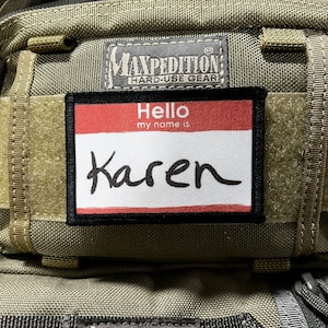 Puede incluir: Una mochila táctica verde y marrón con una etiqueta de nombre negra que dice "Hello my name is Karen". La mochila tiene un parche negro que dice "Maxpedition Hard-Use Gear".