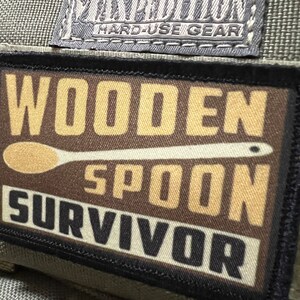 May include: A brown patch with black stitching and the text "WOODEN SPOON SURVIVOR" in white and black lettering. The patch has a Maxpedition Hard-Use Gear logo in the top left corner.