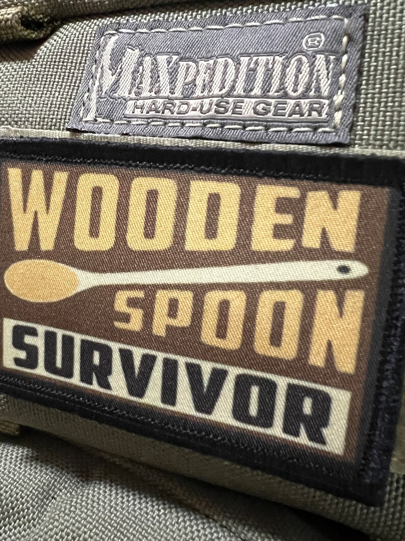 May include: A brown patch with black stitching and the text "WOODEN SPOON SURVIVOR" in white and black lettering. The patch has a Maxpedition Hard-Use Gear logo in the top left corner.
