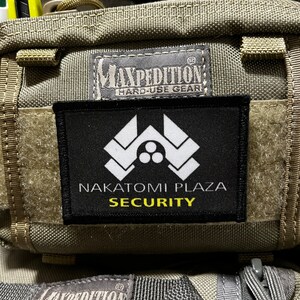 Può includere: Una toppa nera con testo bianco che recita "Nakatomi Plaza Security" con un logo stilizzato bianco sopra il testo. La toppa è attaccata a una borsa verde Maxpedition Hard-Use Gear.