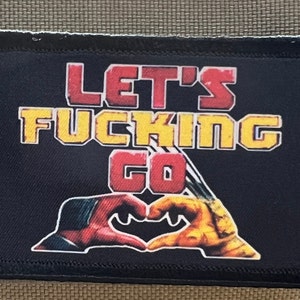 Puede incluir: Parche rectangular negro con las palabras "LET'S FUCKING GO" en letras de bloque rojas y amarillas. Debajo, dos manos forman un corazón, una roja y otra amarilla.
