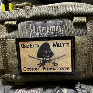 May include: A brown and black patch with a pirate skull and crossbones design. The text "One-Eyed Willy's Custom Booby-Traps" is printed on the patch.