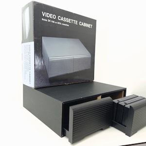 以下が含まれることがあります： 黒いビデオカセットキャビネットと、黒いVHSテープが入った引き出し。キャビネットの後ろには製品の箱が置かれており、白い文字で「VIDEO CASSETTE CABINET」および「Holds 24 VHS or BETA cassettes」と記載されています。