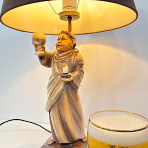 May include: A decorative table lamp with a carved figure holding a chalice and a candle. The lamp has a brown lampshade and a base with a figure in a robe. A glass of beer with foam is next to the lamp.