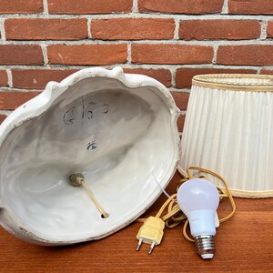 Może przedstawiać: Biała ceramiczna lampa stołowa z białym abażurem z tkaniny. Podstawa lampy ma rzeźbiony wz&oacute;r kobiety trzymającej kwiat. Abażur ma złotą obw&oacute;dkę. Lampa jest włączona i widać żar&oacute;wkę.