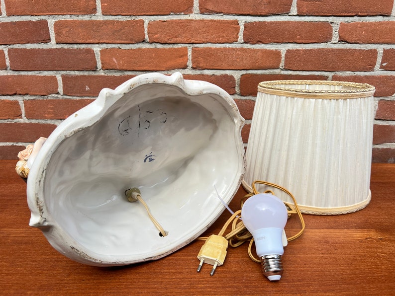 Może przedstawiać: Biała ceramiczna lampa stołowa z białym abażurem z tkaniny. Podstawa lampy ma rzeźbiony wz&oacute;r kobiety trzymającej kwiat. Abażur ma złotą obw&oacute;dkę. Lampa jest włączona i widać żar&oacute;wkę.