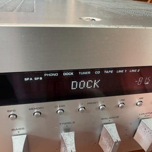 Könnte beinhalten: Ein silberner Audio-Receiver mit einem digitalen Display, das den Text DOCK und -81.5 dB anzeigt. Die Frontplatte enthält verschiedene Bedientasten mit den Beschriftungen PHONO, DOCK, TUNER, CD, TAPE, LINE 1, LINE 2, TP, INFO, MEMORY, CLEAR, PRESET, FM, AM und TUNING.