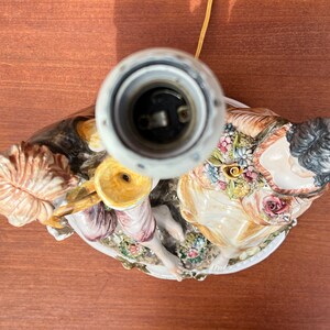 Może przedstawiać: Ceramiczna lampa stołowa z białą podstawą i dwiema postaciami. Postacie to kobieta w kwiecistej sukience i mężczyzna grający na instrumencie muzycznym. Lampa ma złote gniazdo.
