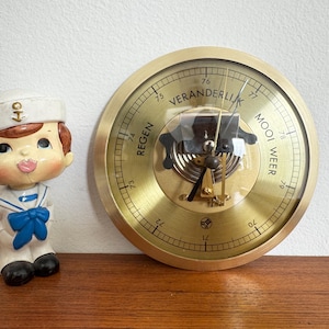 May include: A vintage sailor figurine in a white uniform with a blue bow tie stands next to a brass barometer. The barometer has a glass cover and displays weather indicators in Dutch. The figurine has brown hair and rosy cheeks.
