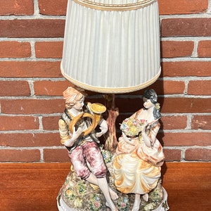 Może przedstawiać: Podstawa lampy wykonana z porcelany przedstawia parę w romantycznym otoczeniu. Mężczyzna gra na rogu, a kobieta trzyma bukiet kwiat&oacute;w. Abażur jest biały z złotą obw&oacute;dką.