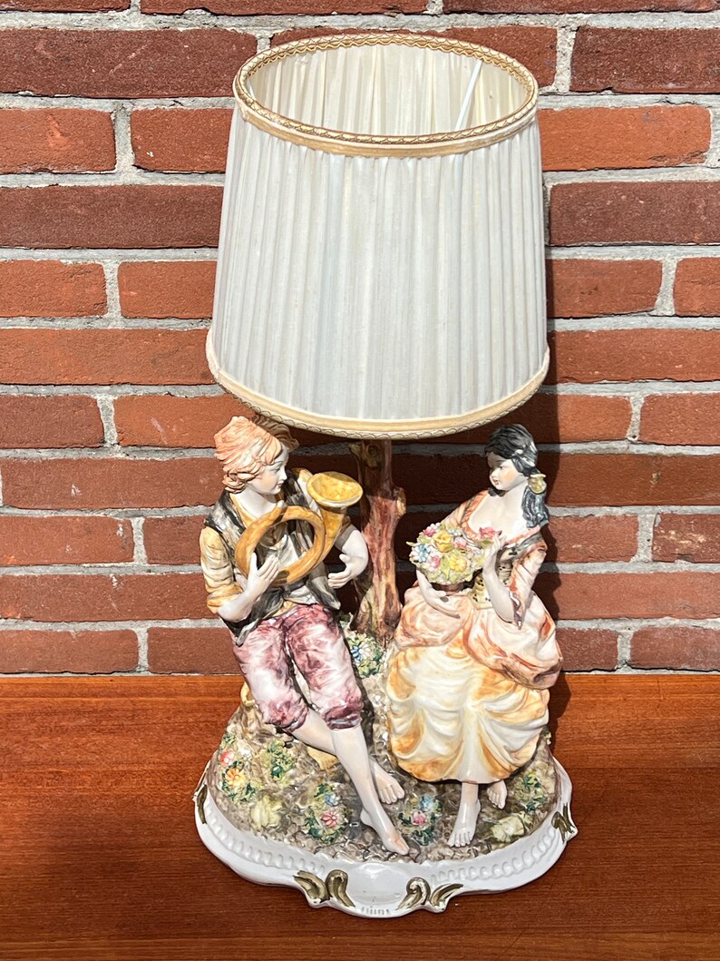 Może przedstawiać: Podstawa lampy wykonana z porcelany przedstawia parę w romantycznym otoczeniu. Mężczyzna gra na rogu, a kobieta trzyma bukiet kwiat&oacute;w. Abażur jest biały z złotą obw&oacute;dką.