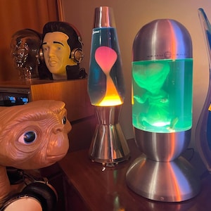 May include: A collection of retro decor items, including two lava lamps with colorful liquid, a sculpture of E.T. the Extra-Terrestrial, and a bust of Elvis Presley wearing headphones. The lamps have silver bases and colorful liquid.