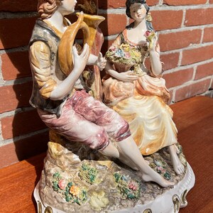 Może przedstawiać: Porcelanowa figurka przedstawiająca parę, mężczyznę grającego na rogu i kobietę trzymającą kosz kwiat&oacute;w. Figurka jest pomalowana w kolorowym stylu z detalami kwiat&oacute;w i liści. Podstawa jest biała z złotą obw&oacute;dką.