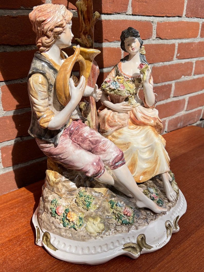 Może przedstawiać: Porcelanowa figurka przedstawiająca parę, mężczyznę grającego na rogu i kobietę trzymającą kosz kwiat&oacute;w. Figurka jest pomalowana w kolorowym stylu z detalami kwiat&oacute;w i liści. Podstawa jest biała z złotą obw&oacute;dką.