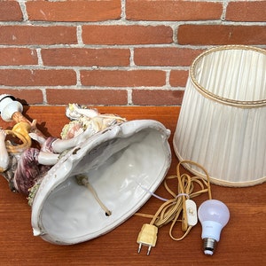 Może przedstawiać: Podstawa lampy wykonana z porcelany z dwoma postaciami grającymi muzykę, białym kloszem i żar&oacute;wką. Podstawa jest ozdobiona kwiatami i liśćmi.