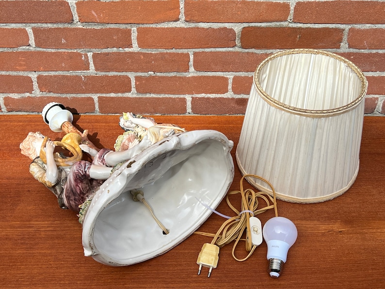 Może przedstawiać: Podstawa lampy wykonana z porcelany z dwoma postaciami grającymi muzykę, białym kloszem i żar&oacute;wką. Podstawa jest ozdobiona kwiatami i liśćmi.