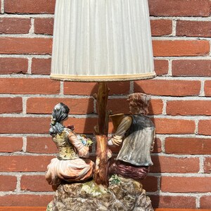 Może przedstawiać: Ceramiczna lampa stołowa z białym abażurem. Podstawa lampy przedstawia parę siedzącą na skalnej formacji, kobieta trzyma kwiaty, a mężczyzna gra na instrumencie muzycznym.