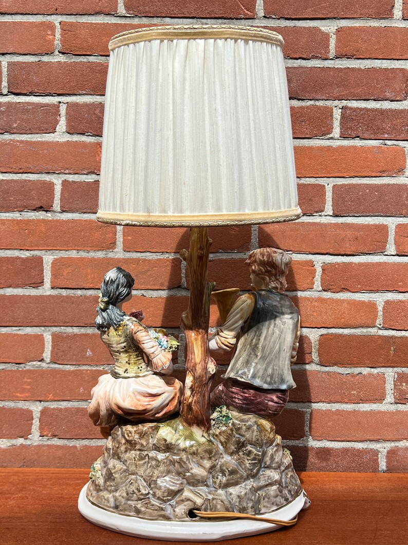 Może przedstawiać: Ceramiczna lampa stołowa z białym abażurem. Podstawa lampy przedstawia parę siedzącą na skalnej formacji, kobieta trzyma kwiaty, a mężczyzna gra na instrumencie muzycznym.