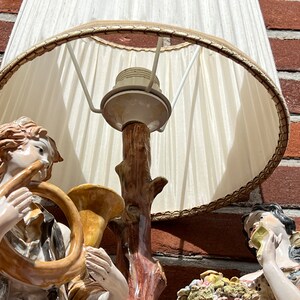 Może przedstawiać: Lampa stołowa w stylu vintage z białym abażurem i porcelanową podstawą przedstawiającą dwie postaci grające na instrumentach muzycznych. Podstawa jest ozdobiona brązowym pniem drzewa i wzorem kwiatowym.