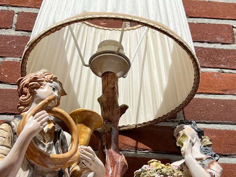 Może przedstawiać: Lampa stołowa w stylu vintage z białym abażurem i porcelanową podstawą przedstawiającą dwie postaci grające na instrumentach muzycznych. Podstawa jest ozdobiona brązowym pniem drzewa i wzorem kwiatowym.