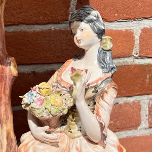 Może przedstawiać: Porcelanowa figurka kobiety w kolorowej sukience trzymającej kosz z kwiatami. Stoi obok pnia drzewa.