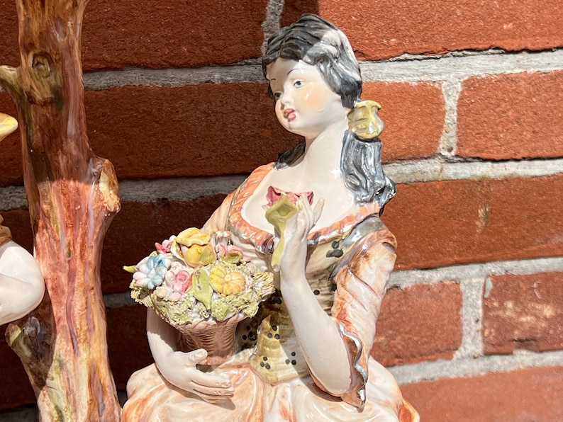 Może przedstawiać: Porcelanowa figurka kobiety w kolorowej sukience trzymającej kosz z kwiatami. Stoi obok pnia drzewa.