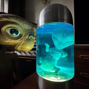 May include: An alien-themed lamp with a blue liquid and a small alien figure inside. The lamp has a silver top with the text "XEMU XENO". An alien head is positioned next to the lamp.