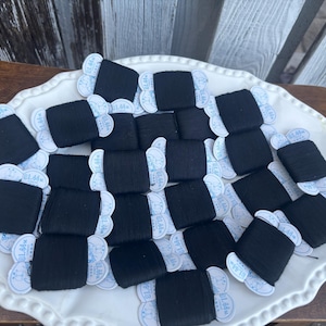 May include: A collection of black thread spools arranged on a white, scalloped-edge serving dish. Each spool is labeled with a small tag. The spools are neatly organized, creating a visually appealing display. The background is a wooden surface.