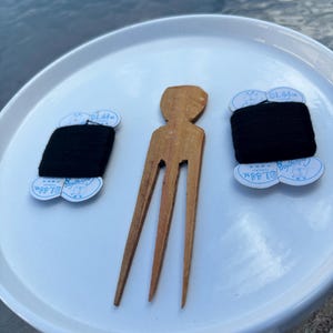 May include: A wooden hair fork and two spools of black thread on a white plate. The thread spools have white labels with blue text. The hair fork is a light brown color.