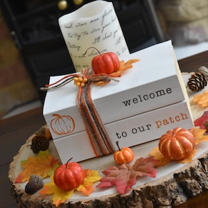 May include: Two stacked white wooden blocks tied with orange and brown twine, featuring the black text "welcome" and "to our patch" with an orange pumpkin outline. The blocks are surrounded by artificial autumn leaves, small pumpkins, and acorns.