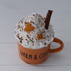 May include: An orange mug with the words "WARM & COSY" in black lettering, filled with whipped cream, cocoa powder, and a cinnamon stick. Two orange leaf decorations are placed on top of the whipped cream. A cozy fall-themed decorative item.