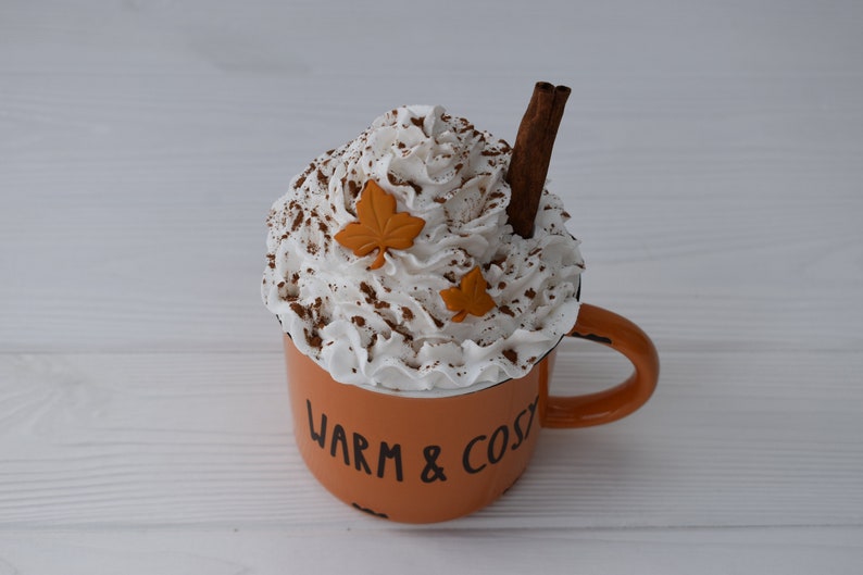 May include: An orange mug with the words "WARM & COSY" in black lettering, filled with whipped cream, cocoa powder, and a cinnamon stick. Two orange leaf decorations are placed on top of the whipped cream. A cozy fall-themed decorative item.
