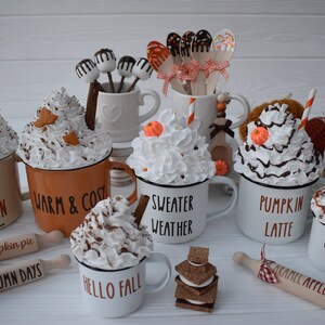 May include: A collection of fall-themed mugs with whipped cream toppings, decorative leaves, and cinnamon sticks. Some mugs have text like "Hello Autumn" and "Sweater Weather." Wooden rolling pins and cake pops are also present.