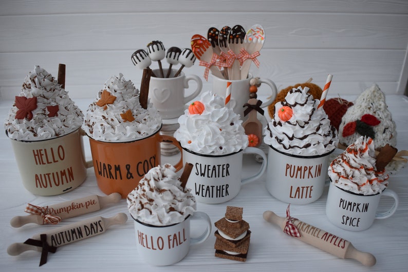 May include: A collection of fall-themed mugs with whipped cream toppings, decorative leaves, and cinnamon sticks. Some mugs have text like "Hello Autumn" and "Sweater Weather." Wooden rolling pins and cake pops are also present.