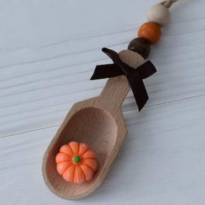 May include: A wooden scoop with a small orange pumpkin decoration inside. The scoop has a dark brown ribbon tied to it, and is attached to a string of wooden beads in various colours. A seasonal autumn-themed craft.