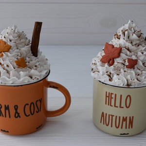 May include: Two decorative mugs with whipped cream topping, cinnamon sticks, and leaf accents. One mug is orange with "WARM & COSY" text, the other is cream-colored with "HELLO AUTUMN" text. Both have black rims.