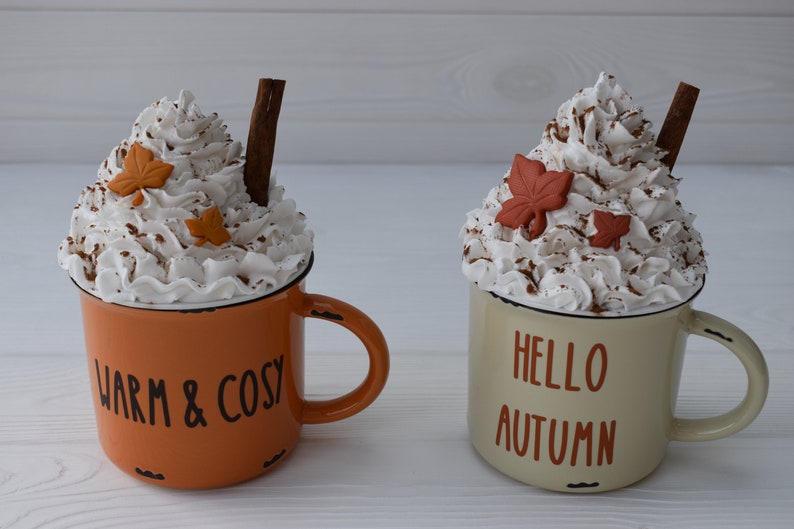 May include: Two decorative mugs with whipped cream topping, cinnamon sticks, and leaf accents. One mug is orange with "WARM & COSY" text, the other is cream-colored with "HELLO AUTUMN" text. Both have black rims.
