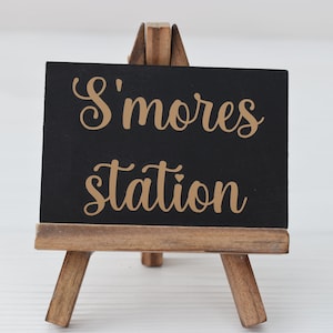 Puede incluir: Un pequeño caballete de madera sostiene una pizarra negra con la inscripción "S'mores station" en letras doradas. El caballete es de madera marrón claro y se encuentra sobre una superficie blanca.