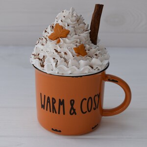 May include: Orange enamel mug with the words "WARM & COSY" in black lettering. The mug is topped with whipped cream, cinnamon, and two leaf-shaped decorations. A cinnamon stick is placed in the whipped cream.