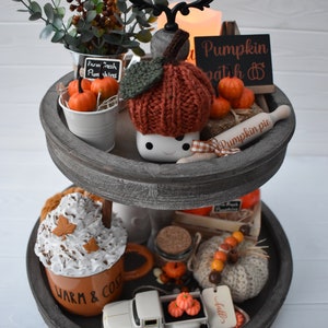 May include: A two-tiered wooden tray with fall-themed decorations. Includes a knitted pumpkin, miniature pumpkins, a mug with whipped cream, and a small white truck carrying pumpkins. The tray is painted gray.