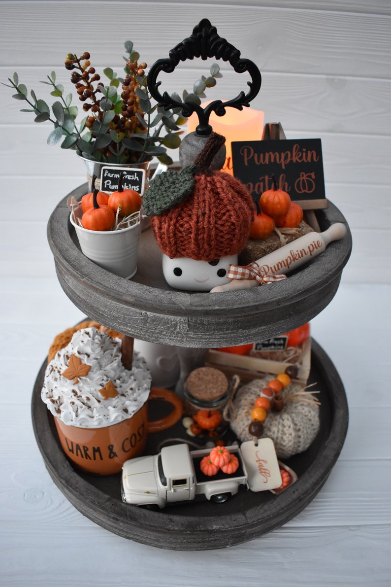 May include: A two-tiered wooden tray with fall-themed decorations. Includes a knitted pumpkin, miniature pumpkins, a mug with whipped cream, and a small white truck carrying pumpkins. The tray is painted gray.