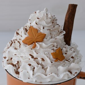 May include: A decorative mug filled with whipped cream, cinnamon, and two orange leaf accents. A cinnamon stick is placed in the whipped cream. The mug is orange with a white rim and a brown handle. The image evokes a fall theme.