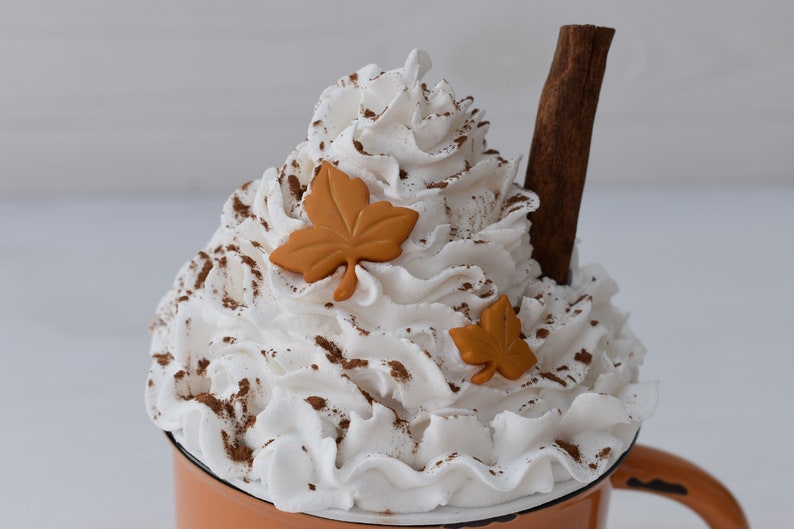 May include: A decorative mug filled with whipped cream, cinnamon, and two orange leaf accents. A cinnamon stick is placed in the whipped cream. The mug is orange with a white rim and a brown handle. The image evokes a fall theme.